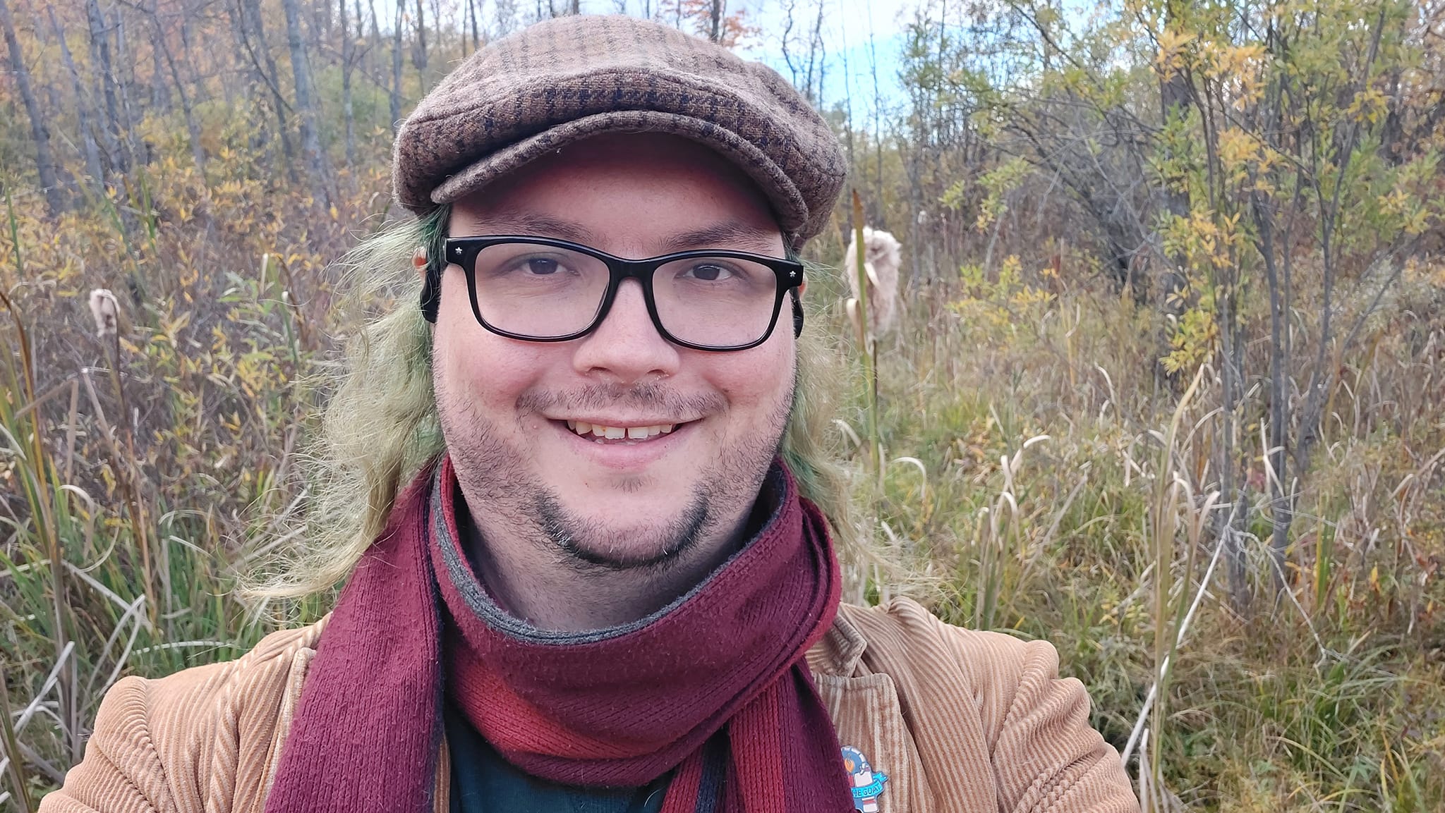 A man with long green hair wearing a green sweater, brown corderoy jacket and red scarf stands in front of some shrubs, most have lost their leaves